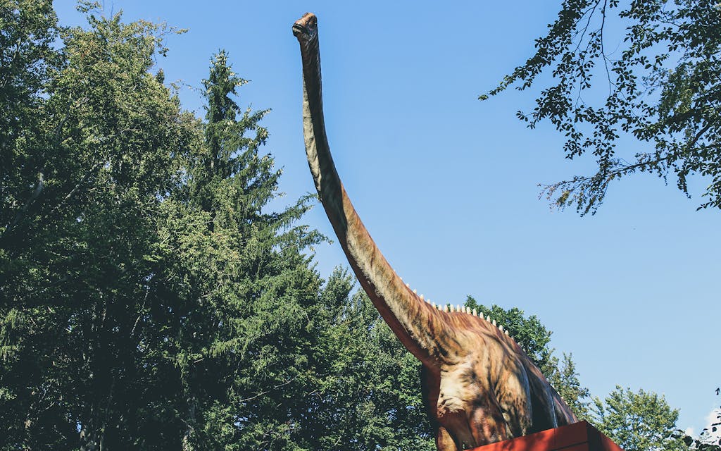 A large dinosaur statue in an outdoor park surrounded by greenery and blue sky.