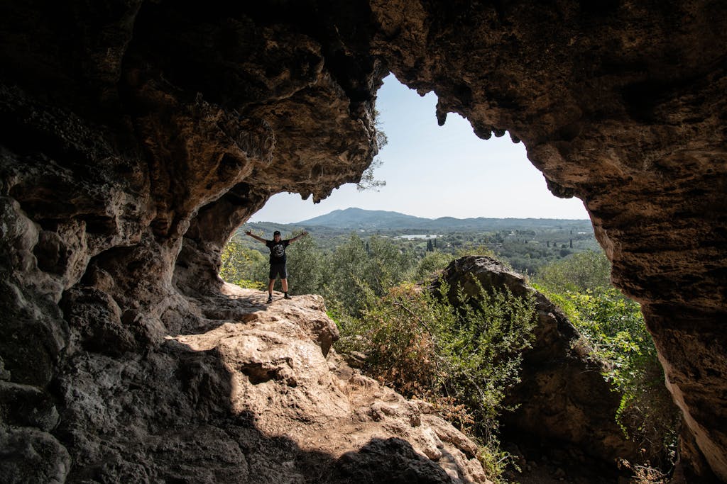 A person stands in a rock cave with scenic views of greenery and mountains beyond.