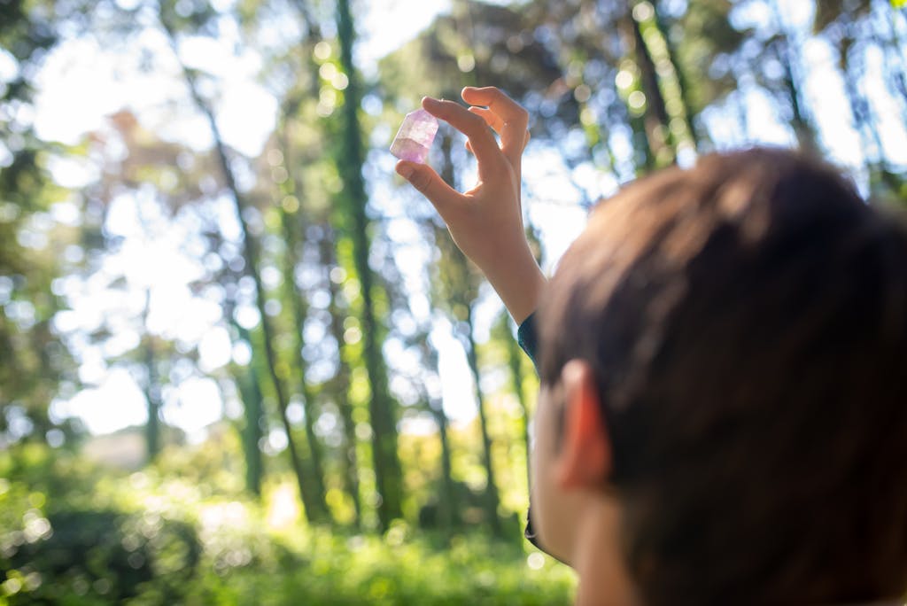 A young individual closely examines a gem under the dappled sunlight in a lush forest setting.