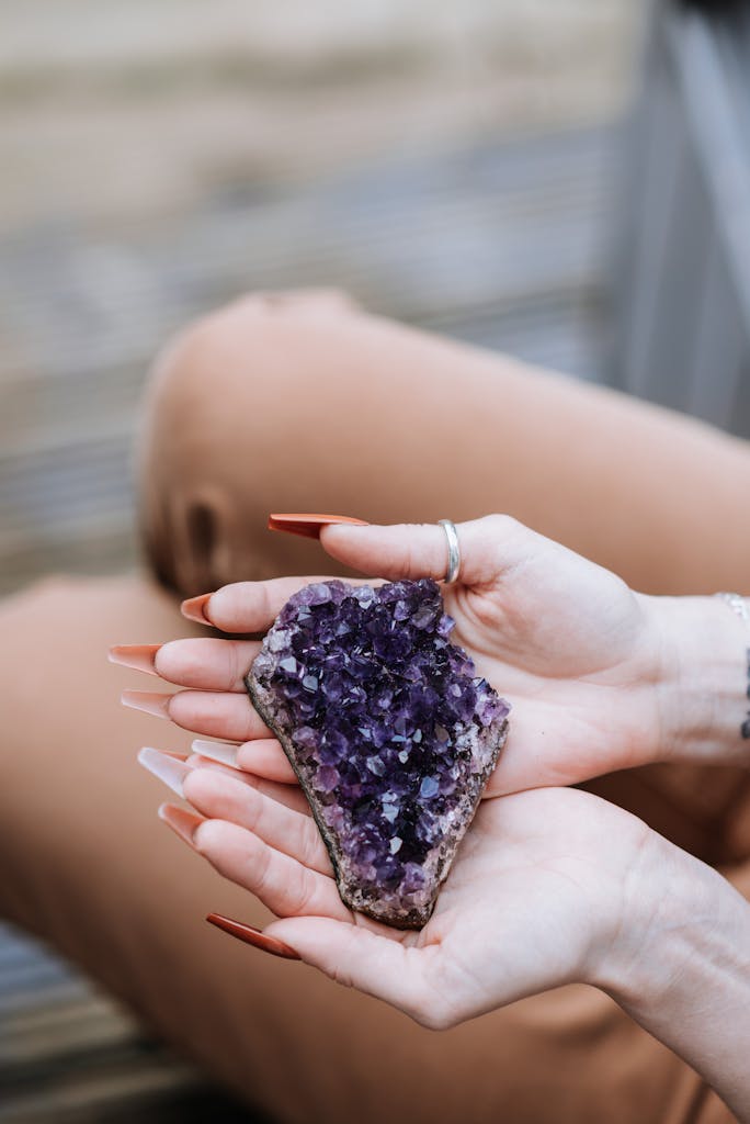 From above of unrecognizable female demonstrating colorful stone with crystal in hands while sitting on wooden steps on blurred background